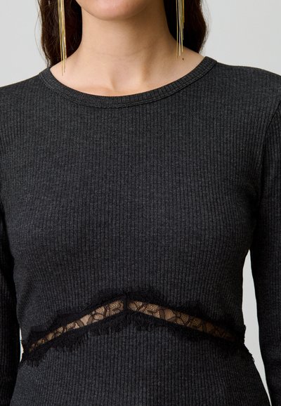 Top a maniche lunghe nero a coste con un pannello in pizzo trasparente con bordo sfilacciato. Texture morbida con scollatura rotonda e vestibilità aderente.