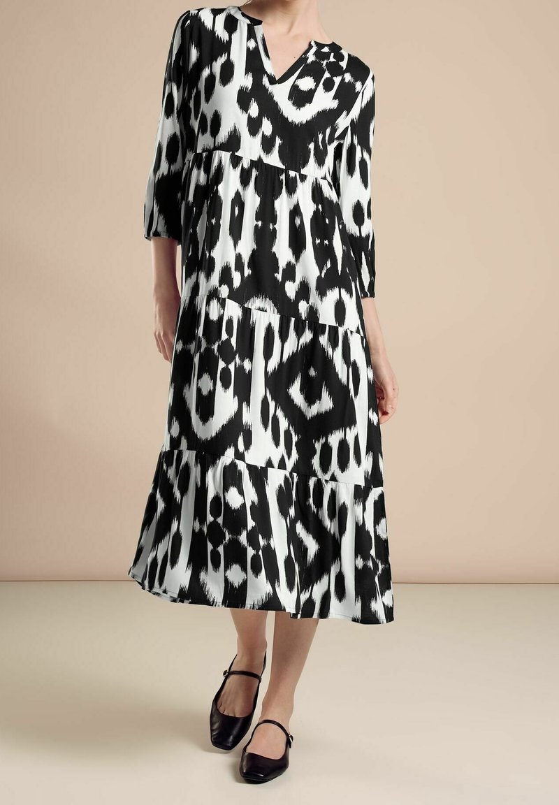 Femme portant une robe midi à imprimé animal noir et blanc avec manches trois-quarts et des chaussures Mary Jane noires, debout devant un fond beige.