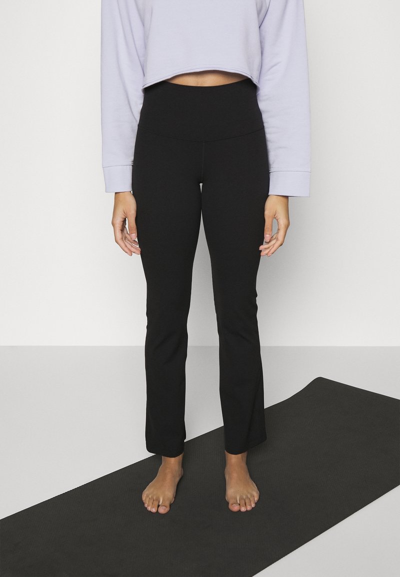 Personne debout pieds nus sur un tapis de yoga noir, portant un pantalon noir taille haute et un haut court à manches longues lavande clair.