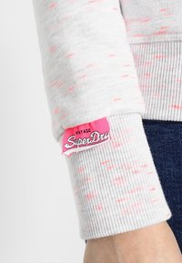 Il polsino di un maglione grigio chiaro presenta una texture a coste con macchie di neon rosa. Include un'etichetta rosa del marchio che dice "Vintage SuperDry."
