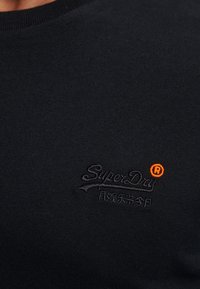 Schwarzes Baumwoll-T-Shirt mit rundem Halsausschnitt und einem dezenten gestickten Logo in dunklem Faden, mit einem orangefarbenen Akzent auf dem "R."