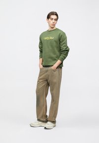 Sudadera verde con texto amarillo "SANTA CRUZ", combinada con pantalones beige. El modelo lleva zapatillas blancas, con un fondo neutro.