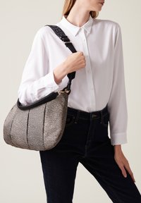 Borsa in tessuto con motivo nero e crema, dalla forma curvata, dotata di una tracolla nera e dettagli in hardware dorato, tenuta contro una camicia bianca.