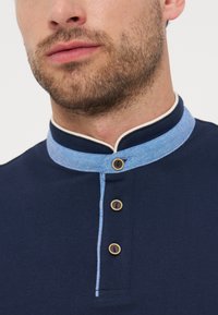 Άνδρας με ελαφρώς μουστάκι που φορά ένα ναυτικό πουκάμισο με ανοιχτό μπλε γιακά μανδαρίνου και τρία ξύλινα κουμπιά στη πατιλέτα.