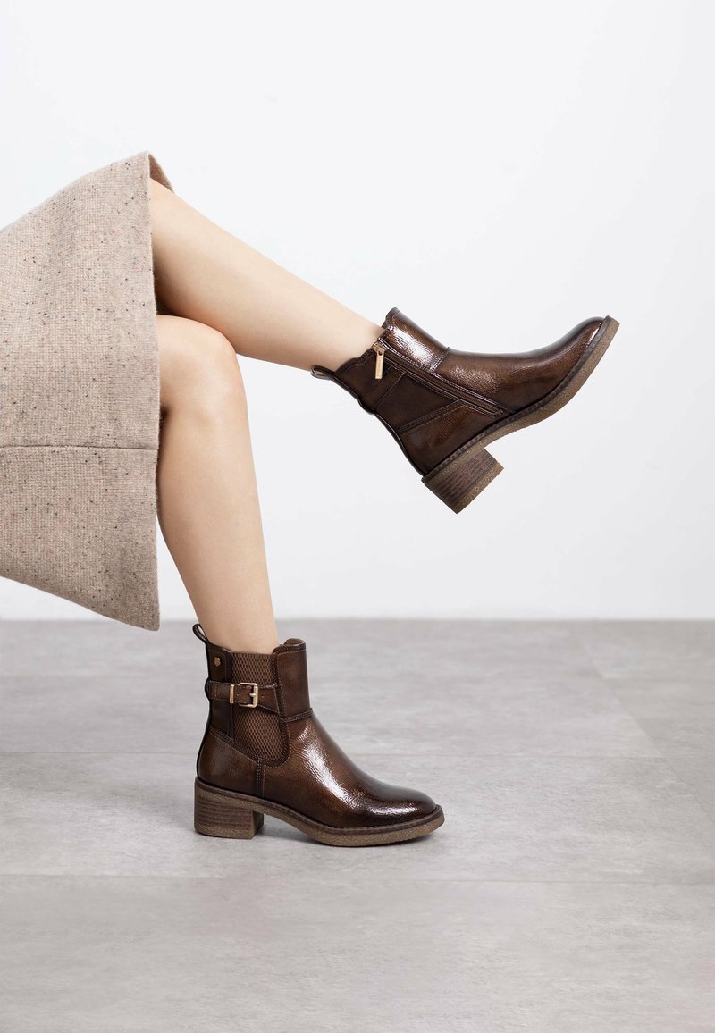 Botas de tobillo de cuero marrón con un acabado texturizado, un tacón bajo y cuadrado, cremallera lateral y una hebilla decorativa en el tobillo.