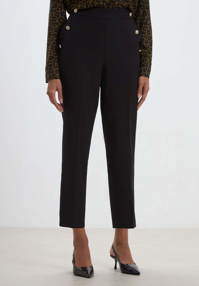 Pantalon noir sur mesure longueur cheville avec boutons dorés décoratifs sur les poches, porté avec des escarpins slingback noirs à bout pointu.