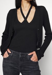 Mujer con un cárdigan negro de canalé sobre una blusa sin mangas negra con tirantes entrecruzados, combinada con jeans de mezclilla gris oscuro, con las manos en los bolsillos.