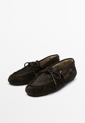 Une paire de mocassins en daim brun foncé avec des bouts ronds et des lacets à nœud sur fond blanc.