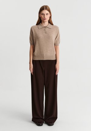 Femme debout de face, portant un polo en maille beige à manches courtes et un pantalon large marron foncé avec des chaussures foncées.