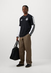 Νεαρός άνδρας που στέκεται, φορώντας αθλητικό μπλουζάκι μαύρο-άσπρο με μακριά μανίκια, καφέ παντελόνι, μαύρα αθλητικά παπούτσια, κρατώντας μια μαύρη τσάντα duffel στο πλάι.