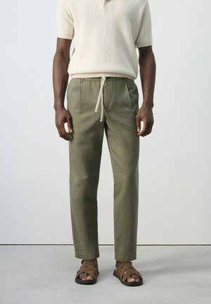 Mężczyzna ubrany w oliwkowe spodnie ze ściągaczem, kremowy krótki sweter z dzianiny i brązowe skórzane sandały, stojący na szarej podłodze.