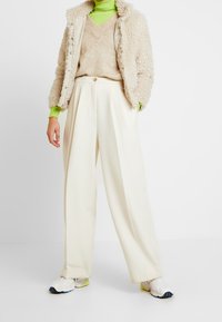 Beige fluffig jacka, krämfärgade vida byxor och grön turtleneck-tröja. Vita och grå atletiska sneakrar. Enkel, modern design.