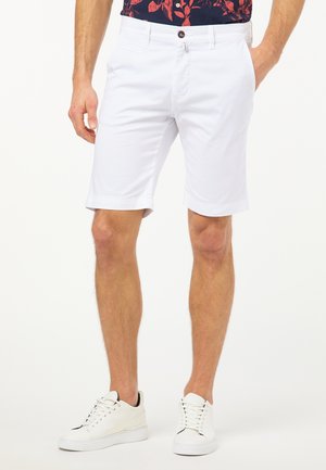 Homme portant un short chino blanc, des baskets blanches et une chemise à fleurs foncée, debout avec une main dans la poche sur un fond blanc.