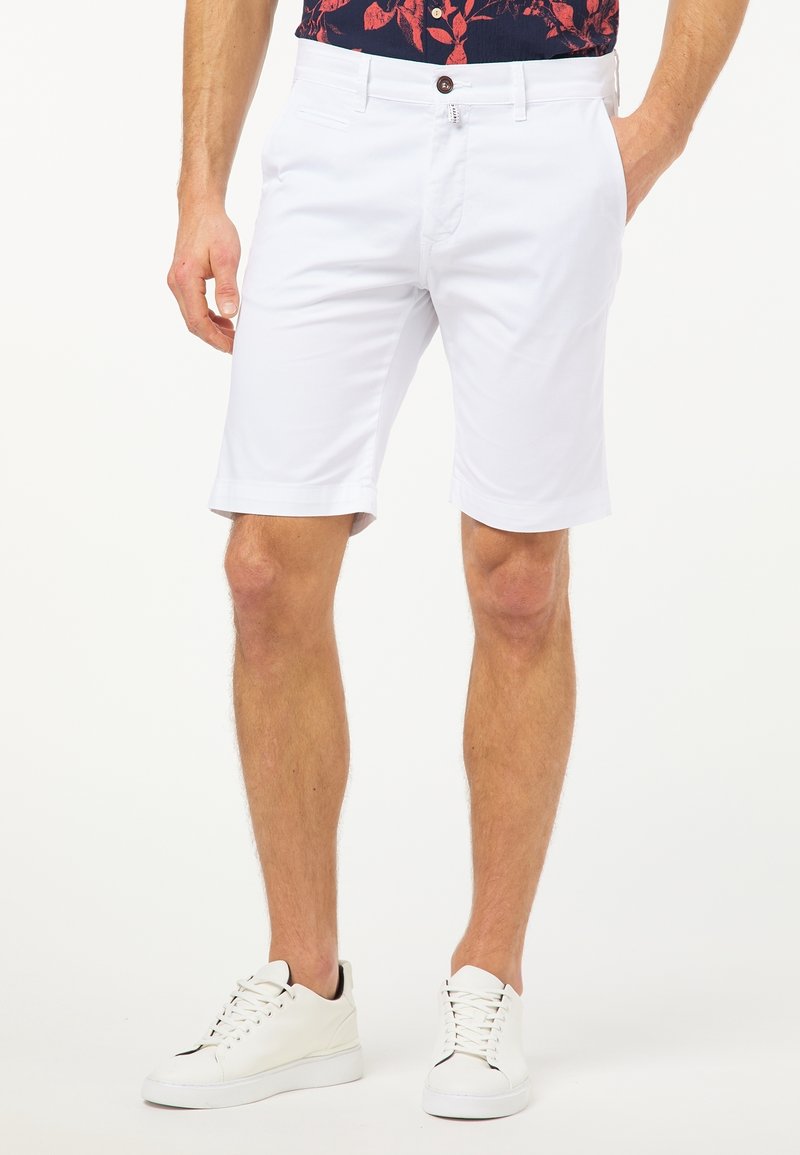 Homme portant un short chino blanc, des baskets blanches et une chemise à fleurs foncée, debout avec une main dans la poche sur un fond blanc.