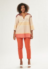 Túnica beige y naranja con escote en V, que presenta rayas horizontales en tonos de marrón, amarillo cremoso y blanco. Combinada con pantalones ajustados naranjas.