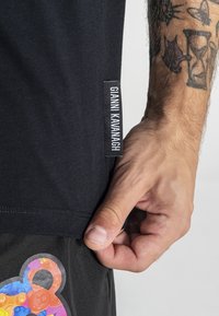 Čierne tričko s postrannou etiketou "Gianni Kavanagh." Tričko má jemnú textúru, kombinované s tmavými nohavicami a farebným akcentovým dizajnom.