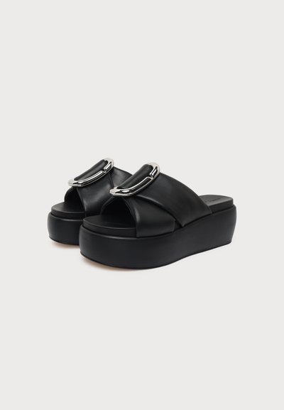 Paire de sandales à plateforme noires avec de larges brides croisées et de grandes boucles argentées sur le dessus, posées sur un fond blanc uni.