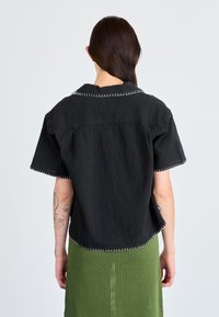Čierna polokošeľa s krátkym rukávom s textúrovanou tkaninou, kontrastným bielym šitím a výstrihom do V, nosená s textured zelenou sukňou.