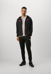 Sort atletisk jakke med røde detaljer og Liverpool FC-logo, parret med sorte tapered bukser og sorte fodboldstøvler. Grå skjorte under.