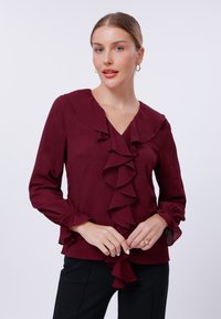 Burgundy blouse with ruffled front and collar, long sleeves with gathered cuffs, soft texture, paired with black pants.