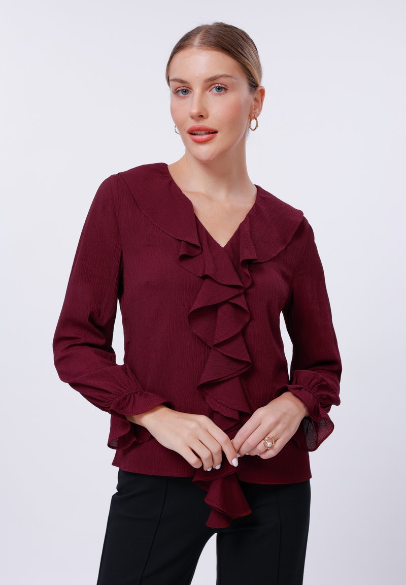 Burgundy blouse with ruffled front and collar, long sleeves with gathered cuffs, soft texture, paired with black pants.