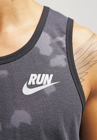 Gros plan sur une personne portant un débardeur camouflé gris foncé avec "RUN" et un logo en forme de virgule imprimés sur la poitrine, ainsi qu'un tatouage géométrique sur l'épaule.