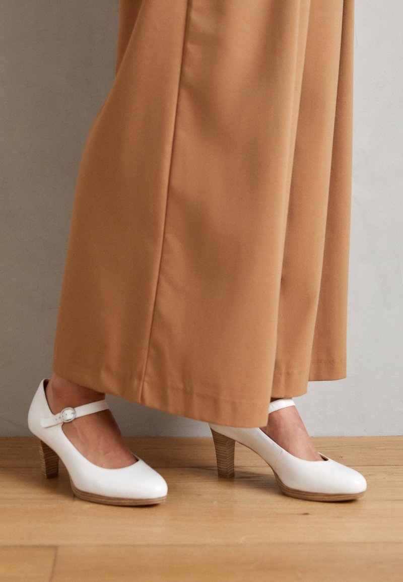 Escarpins Mary Jane blancs en cuir avec un bout arrondi, une bride à la cheville et un talon en bois. Portés avec un pantalon large beige.