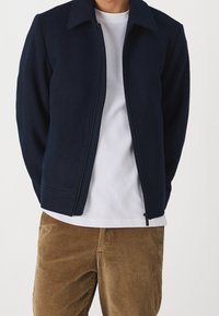Giacca con zip color blu navy e collo, realizzata in un tessuto liscio, con due tasche laterali, indossata sopra una t-shirt bianca e pantaloni in velluto a coste.