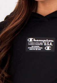 Czarna bluza z kapturem z widoczną etykietą "Champion U.S.A." w białym kolorze i tęczowymi akcentami, wykończona fakturowanym materiałem.