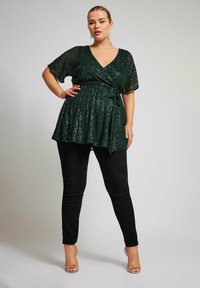 Top avvolgente verde con paillettes, maniche corte, vita aderente e orlo fluido, abbinato a jeans skinny neri e sandali con tacco trasparente.