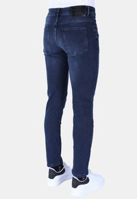 Donkerblauwe slim-fit jeans met een gladde textuur, voorzien van twee achterzakken en een metalen taillebandlabel. Gecombineerd met witte sneakers.