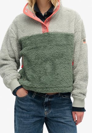 Persoon draagt een tweekleurige groene en lichtgrijze fleece trui met koraalkleurige drukknopen en blauwe spijkerbroek, handen gedeeltelijk in de zakken.