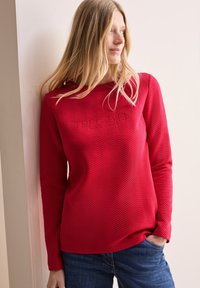 Rode langekorte shirt met een gestructureerd visgraatpatroon en reliëfbranding op de borst. Gecombineerd met blauwe jeans.