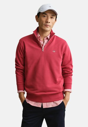 Man met een witte pet, een rood kwart-ritssluitingtrui over een rood-wit gestreept shirt, met handen in de zakken van donkere broek.