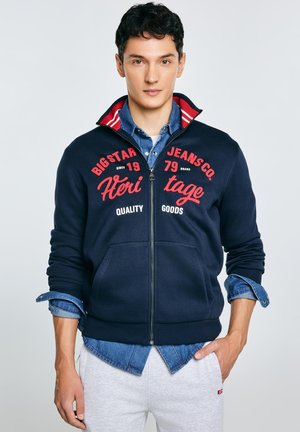 Tmavě modrá mikina na zip s červeným nápisem, bílými detaily a dvěma předními kapsami, vrstvená přes džínovou košili. Hladká textura látky.