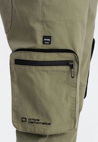 Pantalons cargo vert olive avec une poche zippée, un écusson logo et des détails en métal noir sur le côté. Tissu durable avec une finition texturée.