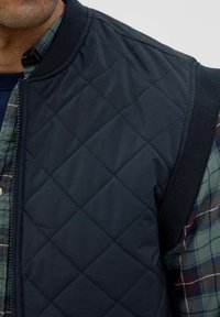 Gilet nero trapuntato con motivo a rombi, dotato di colletto e braccioli a costine, indossato sopra una camicia a quadri verde e blu navy con bottoni visibili.