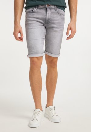 Homme portant un short en jean gris retroussé et des baskets blanches, debout sur un fond uni.