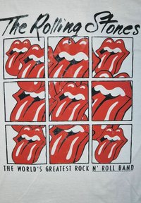 Hvid T-shirt med et grafik af ni røde læber med sorte accenter, en fremtrædende tunge, og teksten "The Rolling Stones" ovenfor og "DEN VERDENS STØRSTE ROCK N’ ROLL BAND" nedenfor.