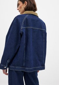 Tumši zila džinsa jaka ar korduroja apkakli. Iespējas ietver brīvu piegriezumu, redzamos šuvumus un pogu akcentus uz aprocēm.