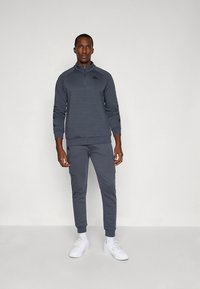 Mörkgrå atletisk träningsdräkt med en dragkedjeförsedd krage och avsmalnande byxor. Slätt tyg med logodetaljer och sidofickor. Vita skor kompletterar outfiten.