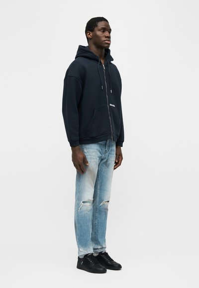 Άνδρας που φοράει σκουρόχρωμο μπλε ζακέτα με φερμουάρ, ανοιχτό μπλε τζιν με σκισίματα και μαύρα αθλητικά παπούτσια, στέκεται μπροστά σε ένα απλό λευκό φόντο.