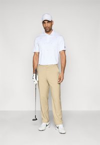 Λευκό πουκάμισο πόλο με μπλε σχέδια, μπεζ παντελόνι και λευκά παπούτσια με γκρι λεπτομέρειες. Κρατάει μια γκολφ κλαμπ, φορώντας λευκό καπέλο και γάντι.