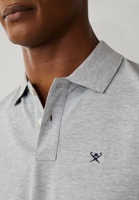 Primo piano di una persona che indossa una polo grigio chiaro con un piccolo logo ricamato raffigurante due mazze da golf incrociate e un cappello sul petto.