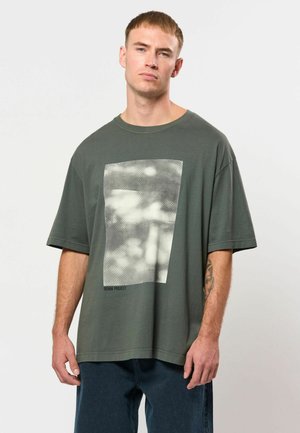 Joven con el pelo corto lleva una camiseta verde oscura de gran tamaño con un gráfico rectangular borroso y vaqueros azul oscuro, de pie frente a un fondo blanco.