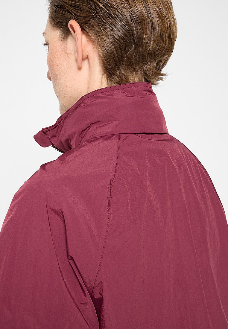 Bordeaux lichtgewicht jack met een hoge kraag en gladde textuur; beschikt over een ritssluiting en minimale stikdetails op de schouders.