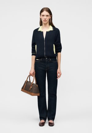 Γυναίκα που στέκεται φορώντας μαύρο φούτερ με φερμουάρ και κίτρινο γιακά, σκούρο τζιν, καφέ παπούτσια και κρατώντας μια καφέ τσάντα σχεδιαστή στο αριστερό της χέρι.