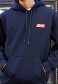 Persona vistiendo una sudadera con capucha de color azul marino con un parche rojo y blanco de "Carhartt Services" en el pecho y las manos en el bolsillo delantero.