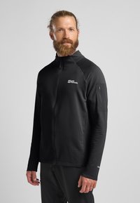 Schwarze Zip-Jacke mit gerippter Textur, hohem Kragen und Logo auf der Brust. Verfügt über eine Reißverschlusstasche am linken Ärmel.