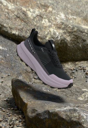 Sort og grå Mammut trail løbesko med tyk sål, der svæver over klippet terræn og viser robust design af udendørs fodtøj.